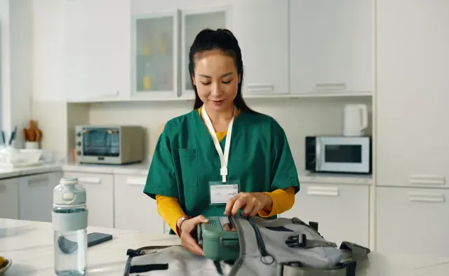 Healthcare professional packing a medical bag while standing in a contemporary kitchen setting with various kitchen appliances visible in background